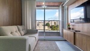 A modern living room with a sofa, a TV mounted on a wooden cabinet, and a sliding glass door leading to a balcony with a city view. - Puerto Ventura #401a - Apto 2 Quartos by Carpediem (João Pessoa)