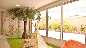 A room with a green artificial grass floor, a beige tent, a large window with a view of a building and plants, and a mural with forest and animal illustrations. - Puerto Ventura #401a - Apto 2 Quartos by Carpediem (João Pessoa)