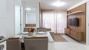 A modern dining area with a white table, cushioned chairs, and a wooden cabinet with a TV. - Puerto Ventura #401a - Apto 2 Quartos by Carpediem (João Pessoa)