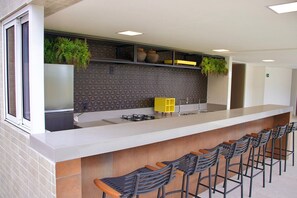 A modern kitchen with a long counter, bar stools, and decorative plants. - Puerto Ventura #305 - Joao Pessoa by Carpediem (João Pessoa)