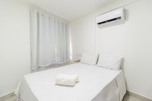 A clean, white hotel room with a neatly made bed, a window with sheer curtains, and an air conditioning unit on the wall. - Puerto Ventura #305 - Joao Pessoa by Carpediem (João Pessoa)