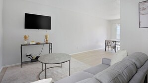 Un salon moderne avec un canapé gris, une table basse ronde, un téléviseur à écran plat fixé au mur et un coin repas à l’arrière-plan.