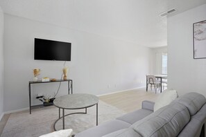 A modern living room with a grey sofa, a round coffee table, a flat-screen TV mounted on the wall, and a dining area in the background. - Updated 2BR 2BA with full kitchen (San Antonio)