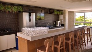 A modern kitchen with a bar area, featuring a countertop with glasses, a built-in stove, and a large window offering a view of the city. - Puerto Ventura #303 - Flat by the sea in Carpediem (João Pessoa)