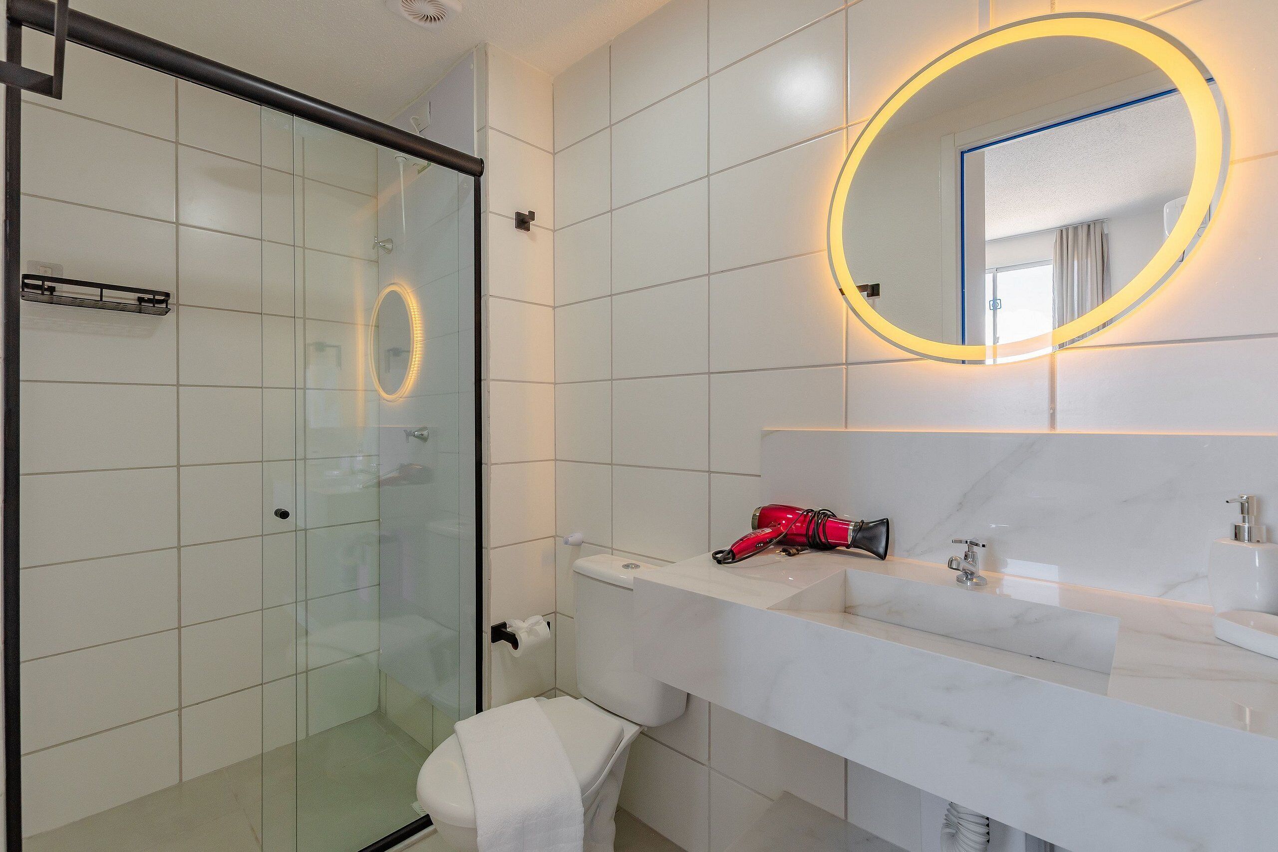 Un baño moderno con cabina de ducha de vidrio, un lavamanos blanco con encimera de mármol y un espejo redondo con luz amarilla.