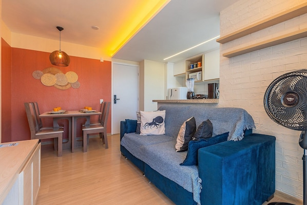 A modern living room with a blue sofa, wooden dining table, and chairs. There is a kitchen area with a microwave and a fan. - Mirante #1401 - Complete Apartment by Carpediem (Fortaleza)