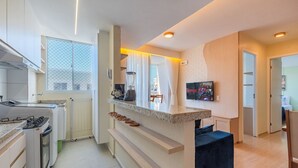 Une cuisine moderne avec des meubles blancs, un four encastré et un plan de travail équipé d’un mixeur. On y trouve une fenêtre avec des rideaux légers, un petit coin salon avec un banc et un téléviseur fixé au mur.