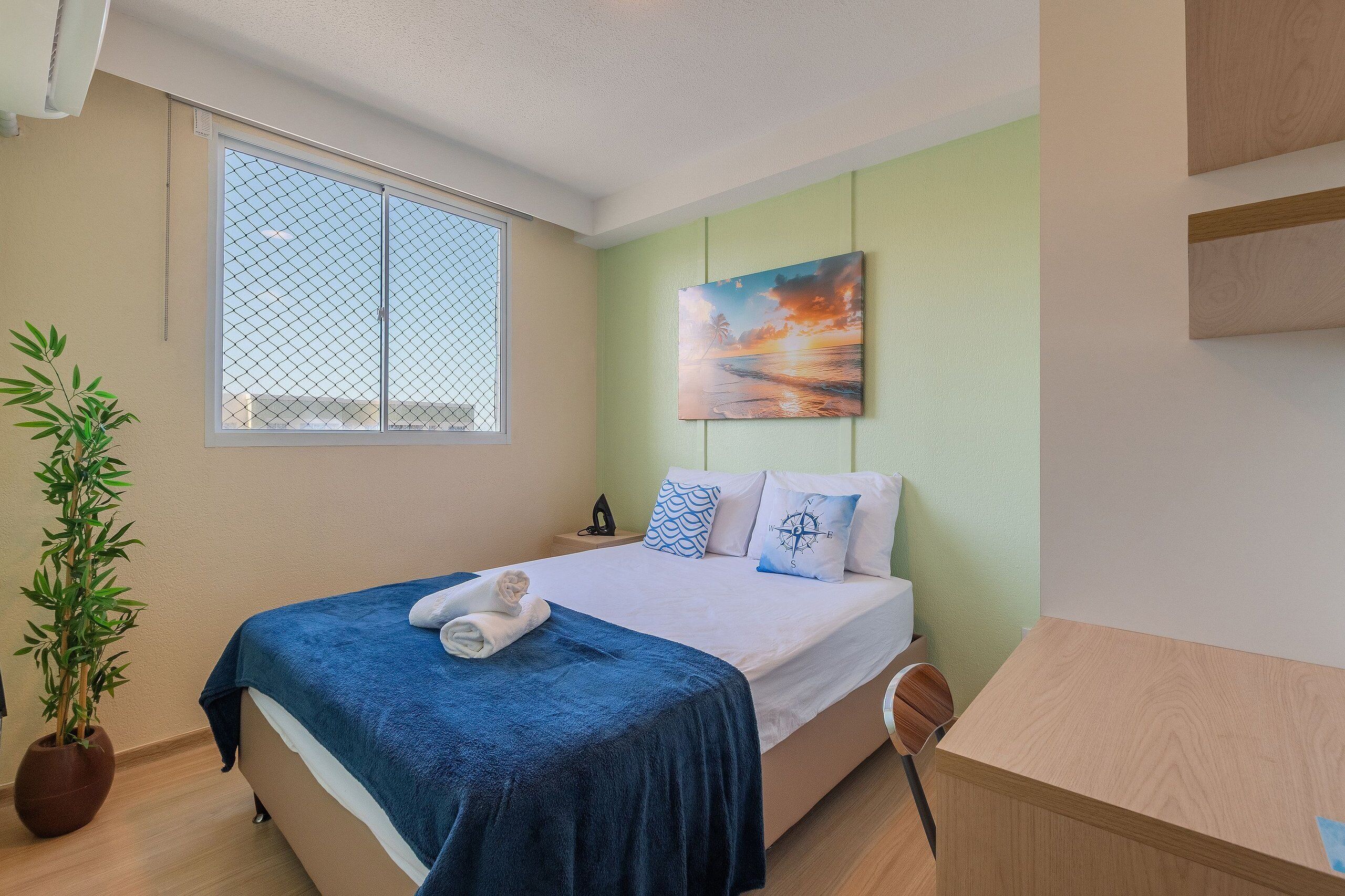Una cama bien hecha con un edredón azul, una planta en maceta y un cuadro de un atardecer en la pared.