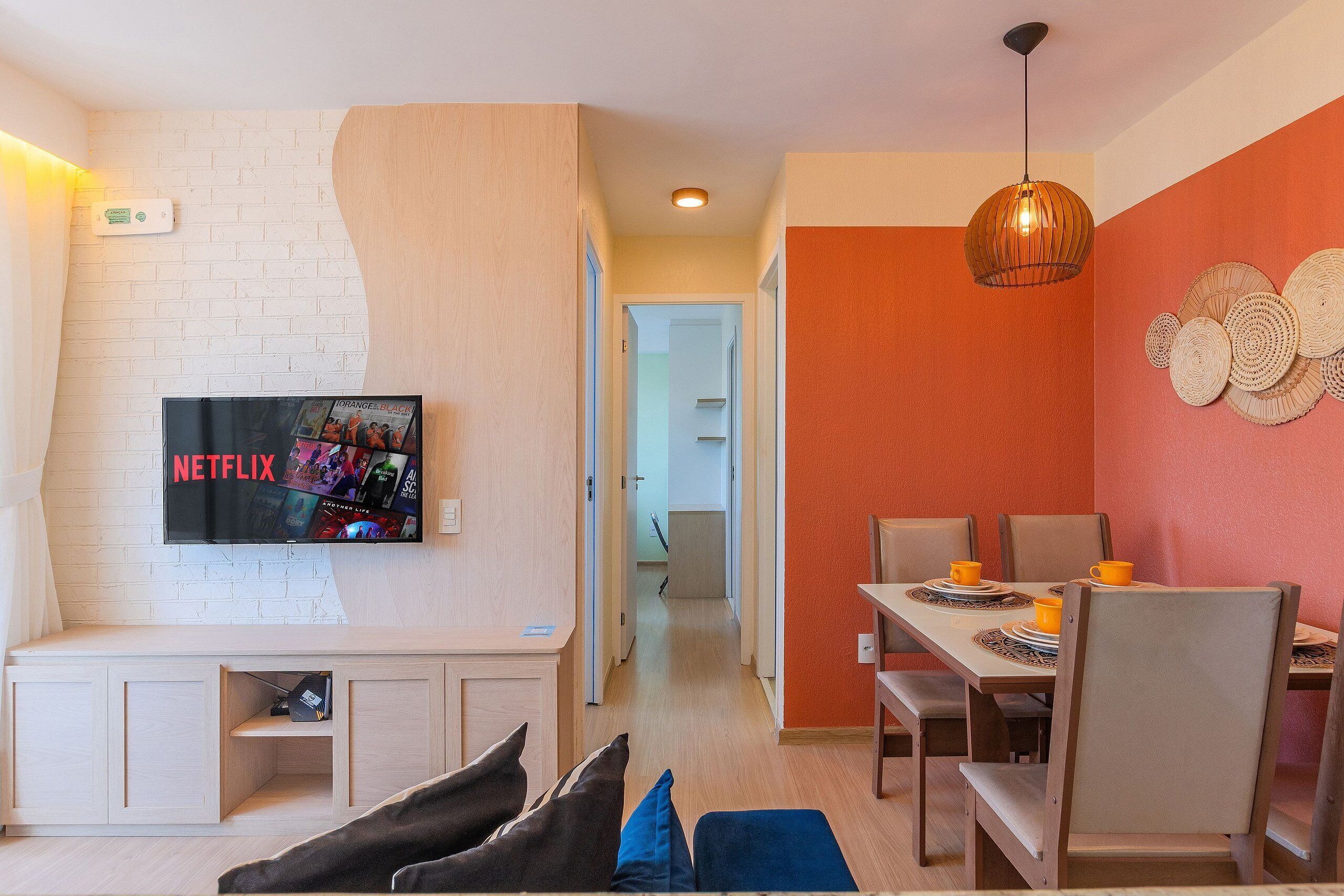 Uma sala de estar moderna com televisão de tela plana, um espaço de refeições com mesa e cadeiras, e um corredor que dá acesso a outros cômodos.