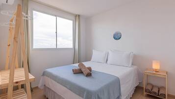 Un dormitorio con una cama, una silla de madera, una mesita de noche con lámpara y una ventana con cortinas.