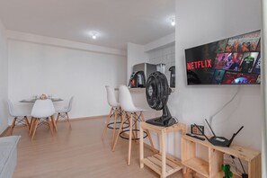 Ein modernes Wohnzimmer mit Essbereich, einem Standventilator, einem Fernseher, der Netflix zeigt, und einem kleinen Holztisch mit Laptop und Fernbedienung.