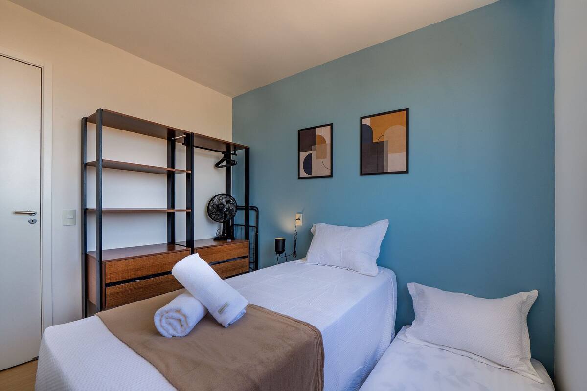 A hotel room with a bed, a wooden nightstand, a fan, and two abstract paintings on the wall.
