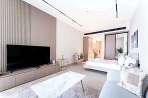 A modern living room with a large flat-screen TV, a white sofa, a coffee table, and a dining area with a geometric glass table. - Stayshort - Amazing Studio in JVC That Sleeps 2 With Balcony (Dubai)