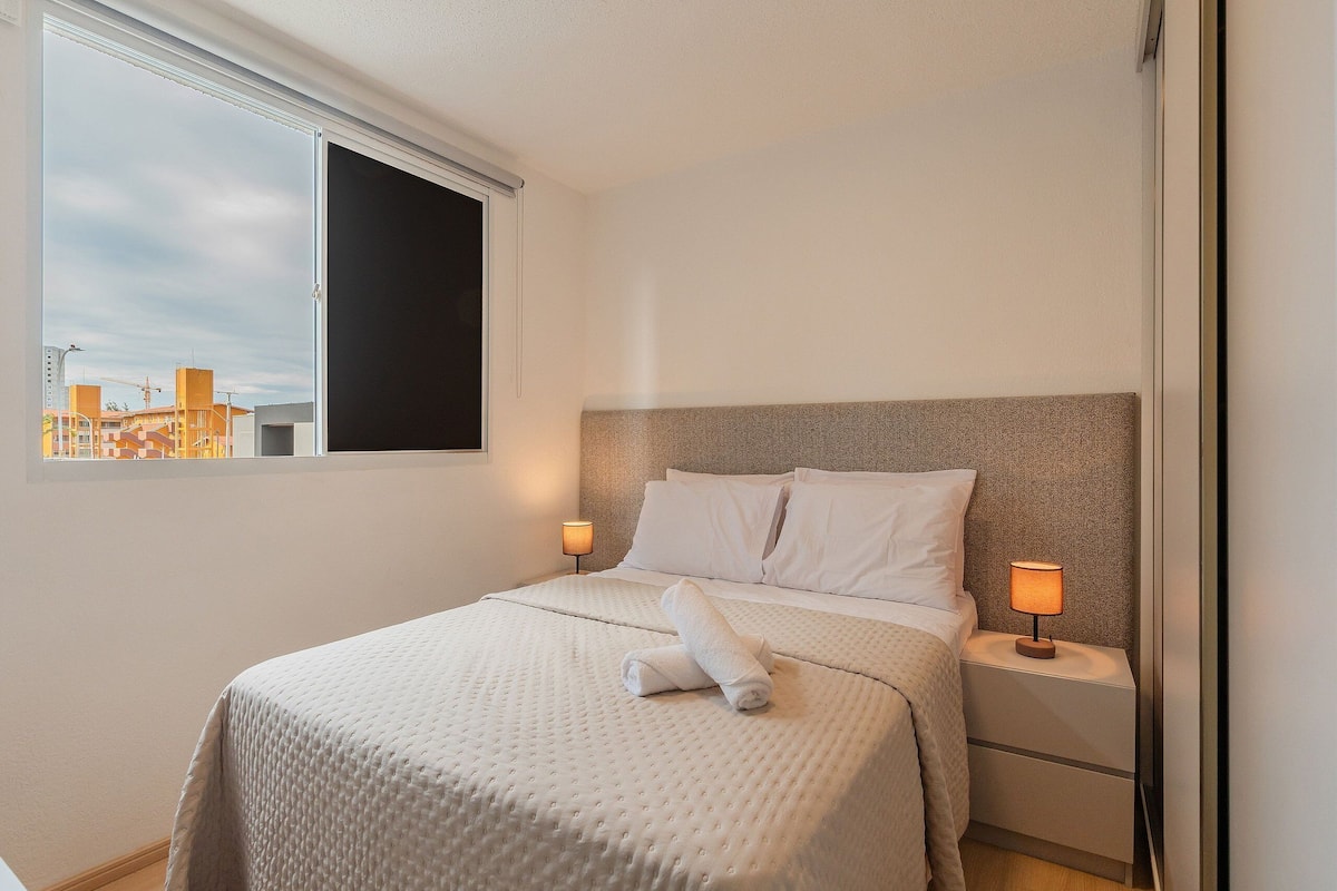 Una cama bien hecha con edredón blanco y dos almohadas, una mesita de noche con lámpara, y una ventana con vistas a edificios.