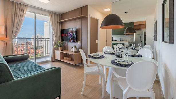 Uma sala de estar moderna com um sofá verde, uma mesa de jantar posta para quatro pessoas e uma cozinha ao fundo.