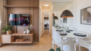 Uma sala de estar moderna com uma televisão de tela plana, uma área de jantar com cadeiras brancas e uma mesa, e um corredor que dá para outro quarto.