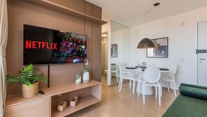 Uma sala de estar moderna com televisão de tela plana, uma área de jantar com cadeiras brancas e um sofá verde.