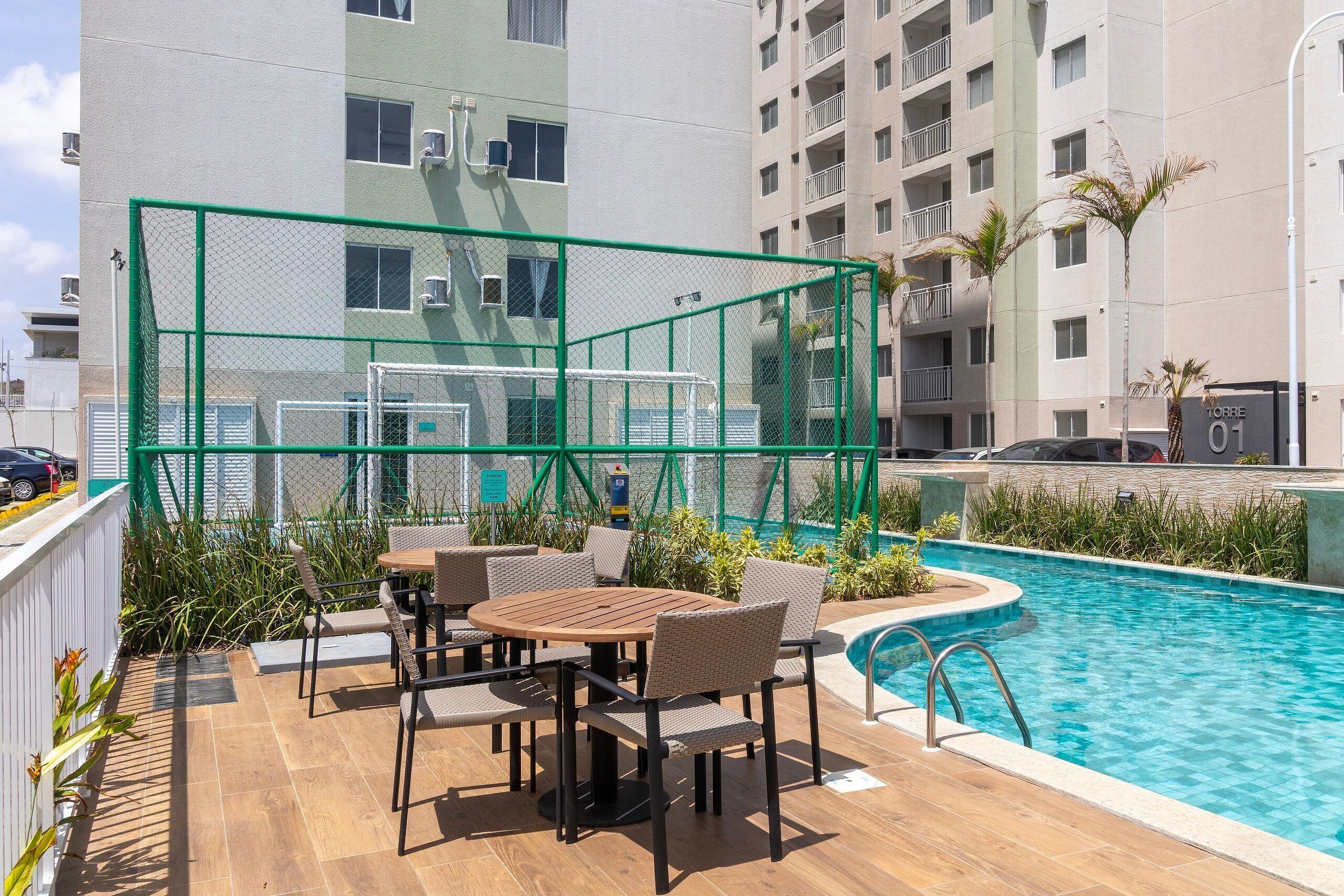 Uma área à beira da piscina com mesa e cadeiras de madeira, cercada por uma cerca verde.