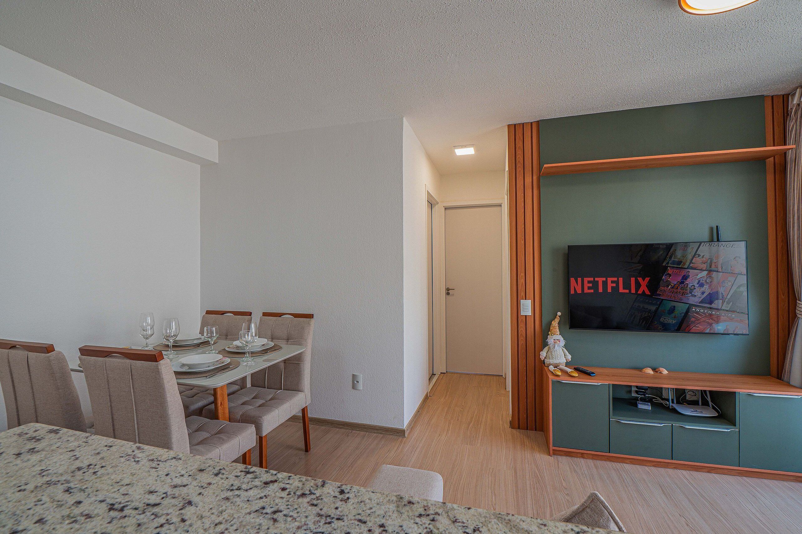 A modern living room with a dining area, a flat-screen TV mounted on a wooden wall, and a door leading to another room.