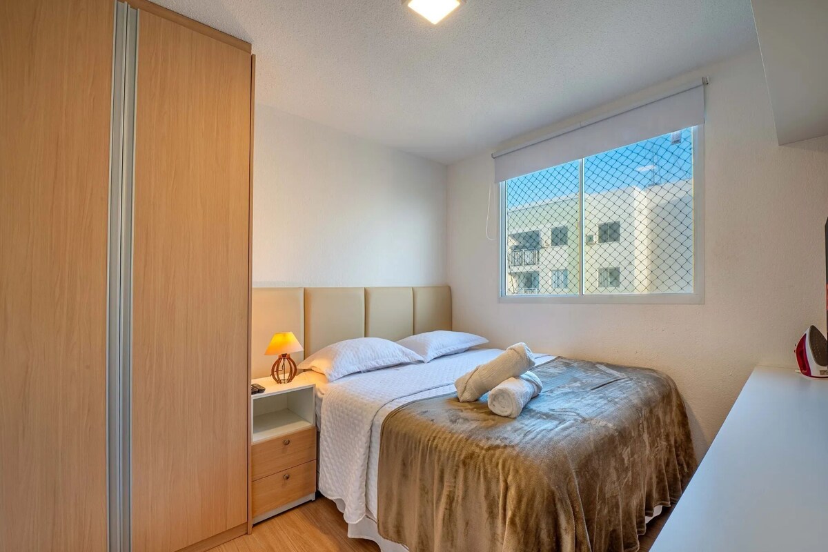 A bedroom with a bed, a nightstand, a lamp, a window with a view of buildings, and a wardrobe.