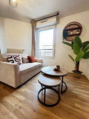 A living room with a sofa, two round tables, a potted plant, and a window with a view. - Modern Apartment Near Paris & Orly • Pvt Parking (Athis-Mons)