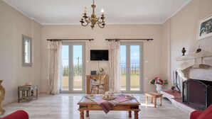 A living room with a fireplace, a television, a table with a placemat, and a balcony with curtains. - Creta Leisure Escape, Private Pool & Garden Oasis (Pompia)