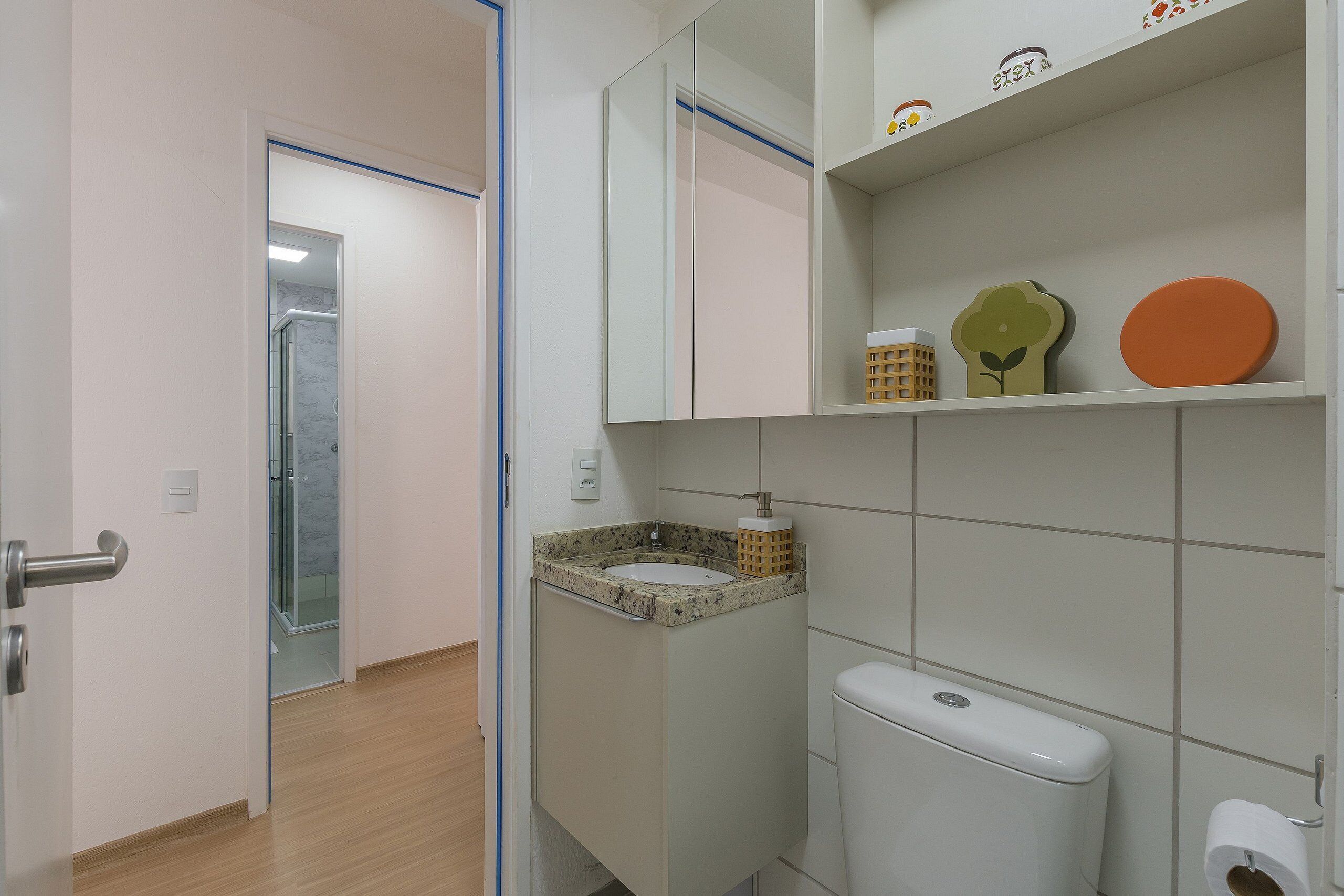 A bathroom with a white sink, a toilet, and a mirror above the sink. There are shelves with decorative items and a door leading to another room.