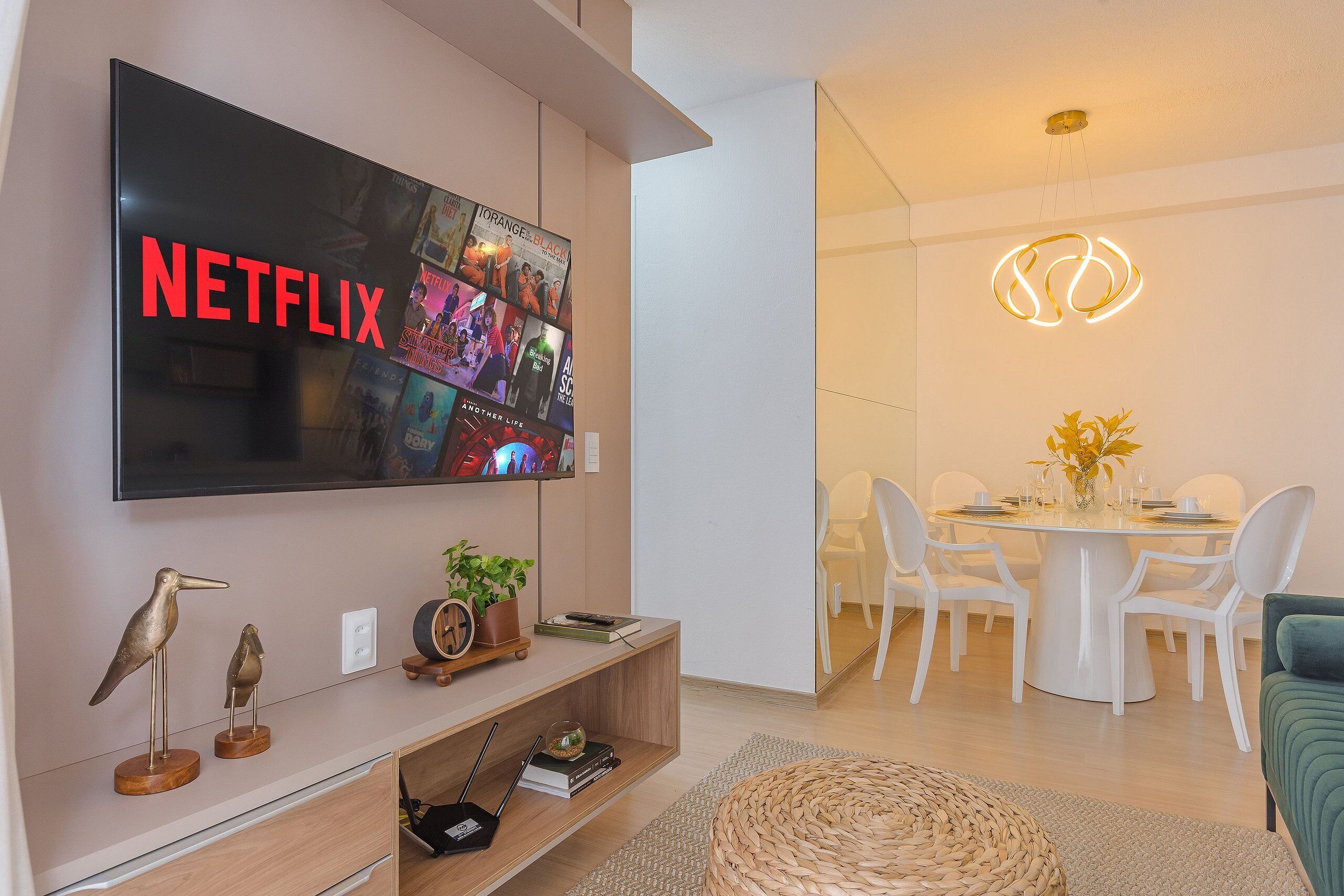 Un salon moderne doté d’un grand téléviseur à écran plat affichant le logo de Netflix, d’un coin repas avec une table et des chaises blanches, et d’un luminaire au plafond au design original.