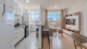 A modern apartment with a kitchen, dining area, and living room. It features a large TV, a balcony with a view, and a sliding door to the outside. - Mirante #303 2 Suites Praia Futuro by Carpediem (Fortaleza)