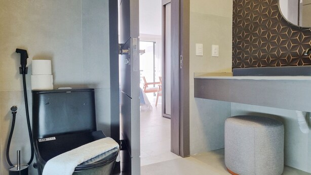 A modern bathroom with a black toilet, a towel, a grey ottoman, and a mirror with a geometric pattern. - Iate Plaza #1404 - Studio Vista Mar por Carpediem (Fortaleza)