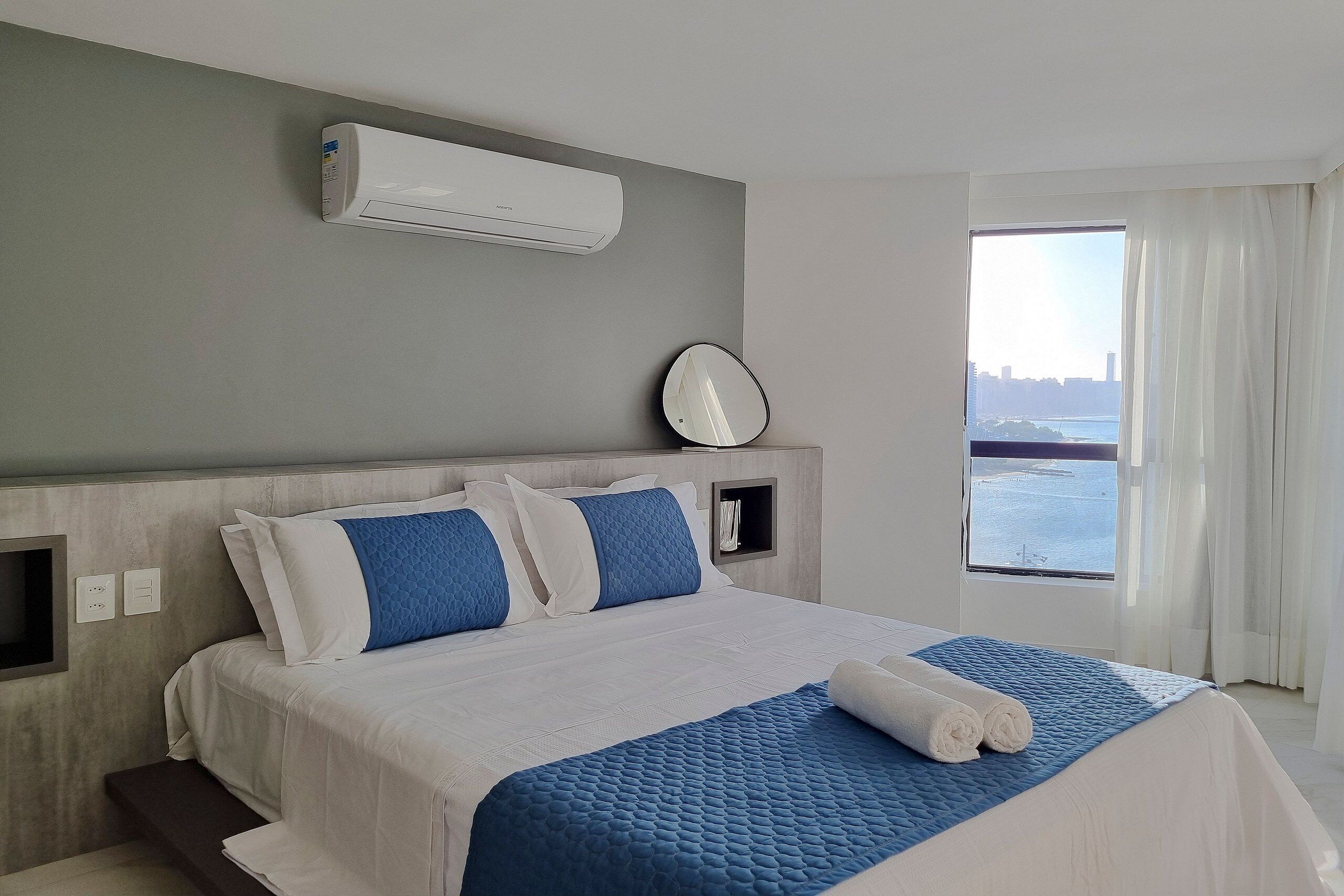 A hotel room with a bed, a window with curtains, an air conditioning unit, and a wall-mounted clock.