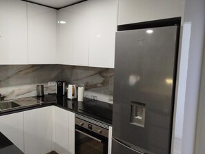 Una cuina moderna amb armaris blancs, una barra de resguard de marbre, un frigorífic d'acer inoxidable i un forn integrat.