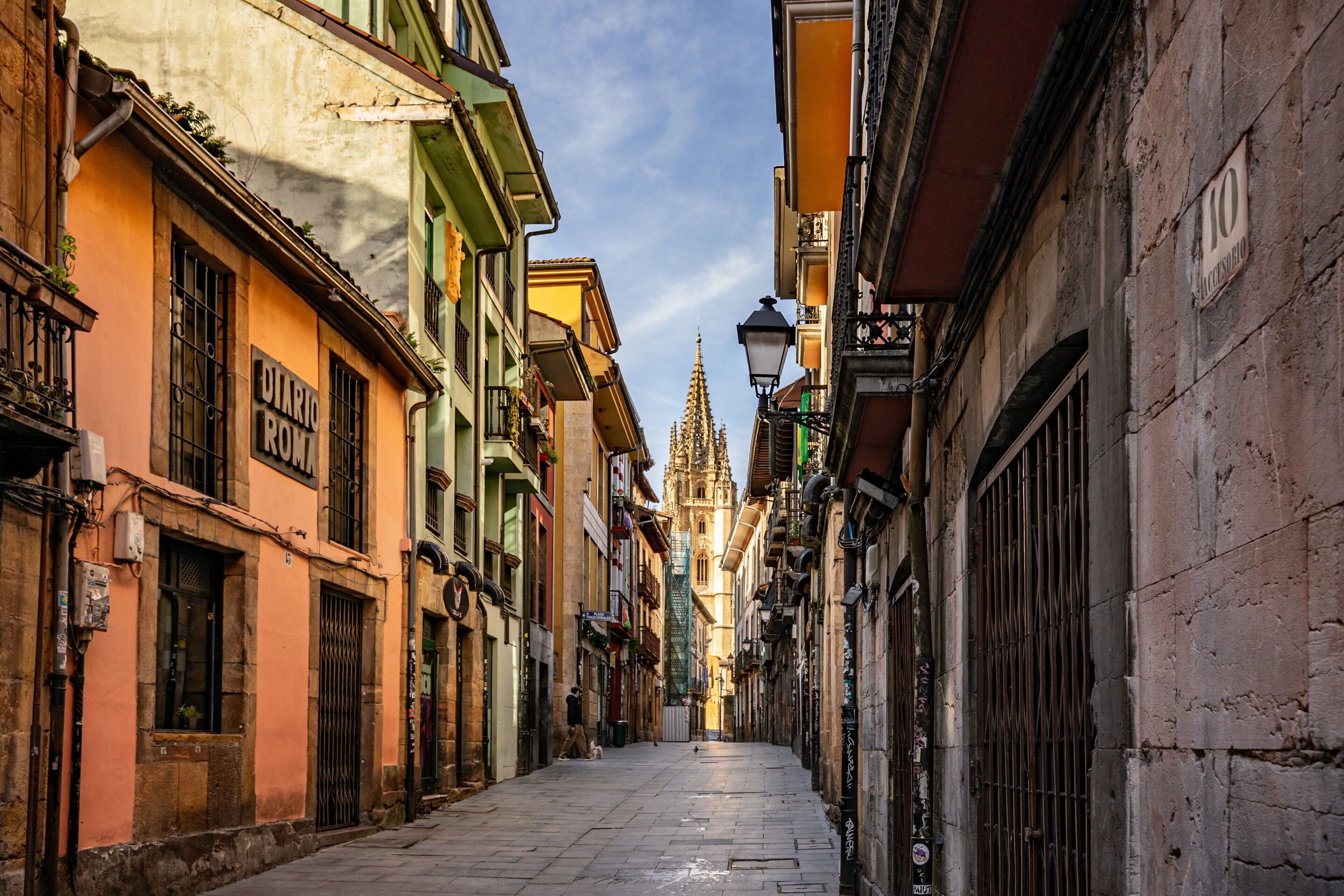 A narrow street with historic buildings, a cathedral spire, and a street lamp.