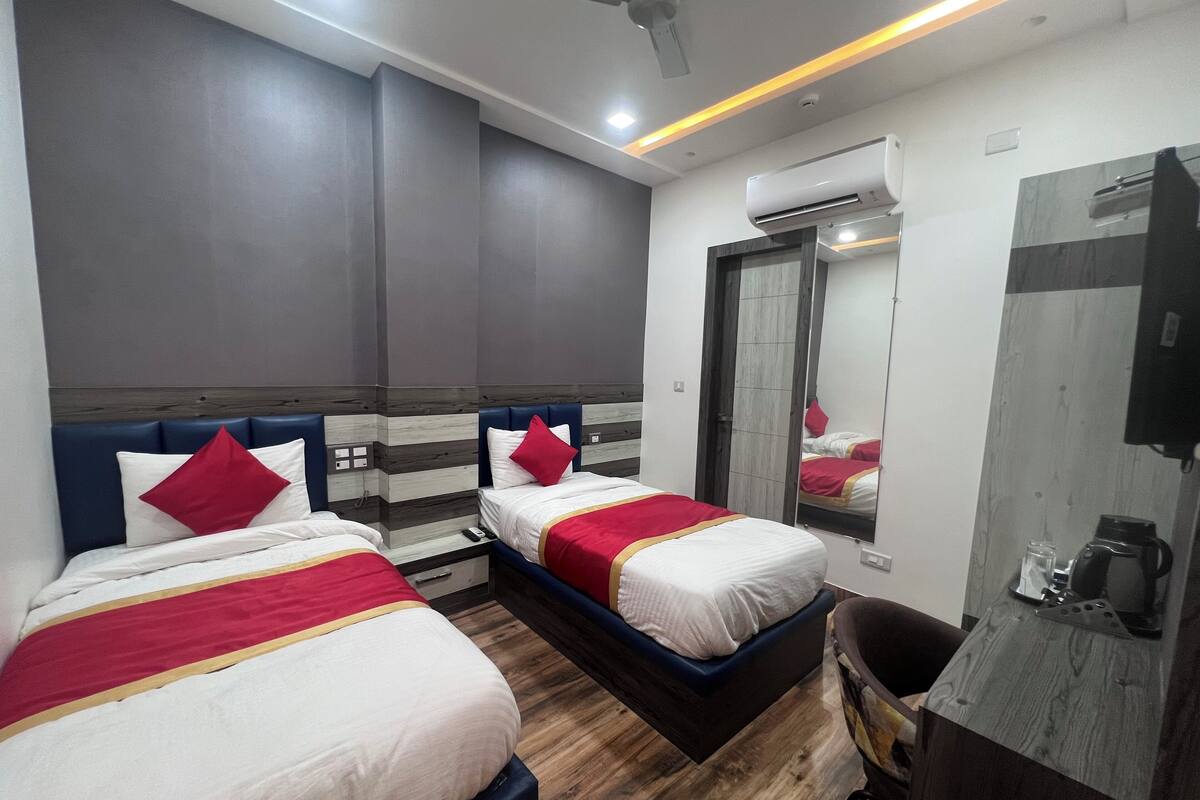 Habitación de hotel con dos camas, cada una con cubrecama rojo y blanco, suelo de madera, ventilador de techo, aire acondicionado y un espejo en la pared.