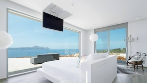 Una camera da letto moderna con un letto grande, una TV montata a soffitto e vista mare attraverso ampie finestre.