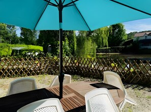An outdoor patio with a table and chairs, a blue umbrella, and a view of a river and greenery. - Holiday Home 'Moulin De La Forge' with Lake View, Private Pool and Wi-Fi (Les Arques)