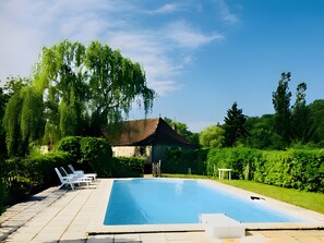 Une piscine entourée de chaises longues, de verdure et d’un bâtiment dont le toit est recouvert de tuiles.