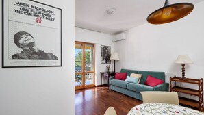 Una habitación con un sofá verde, una cama con colcha floral, una mesita de noche de madera, una lámpara colgante y un cartel en la pared.