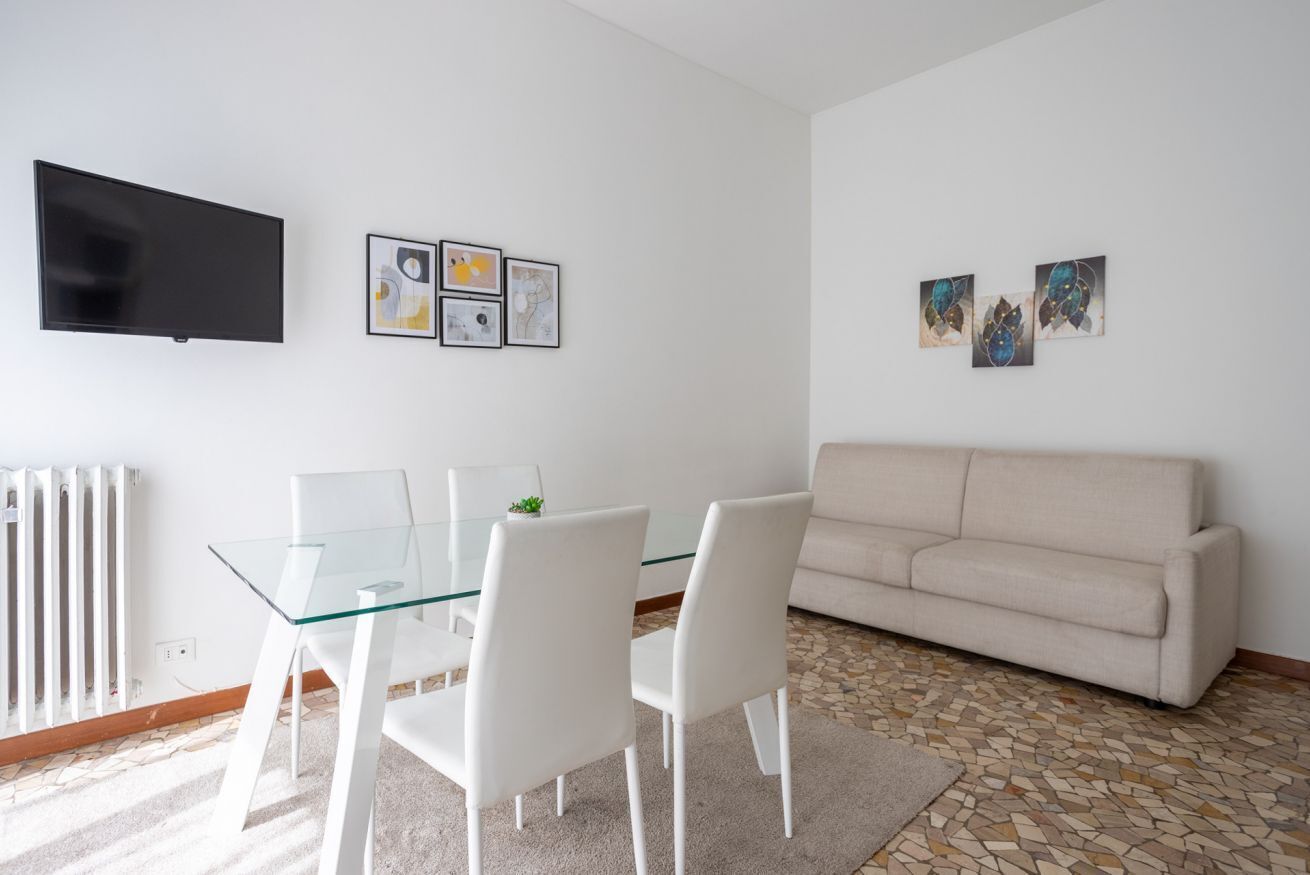 Un salon moderne avec une table à manger en verre, des chaises blanches, un canapé beige, un téléviseur à écran plat et des œuvres d’art abstraites accrochées aux murs.