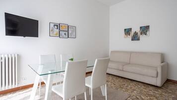 Un salon moderne avec une table à manger en verre, des chaises blanches, un canapé beige, un téléviseur à écran plat et des œuvres d’art abstraites accrochées aux murs.