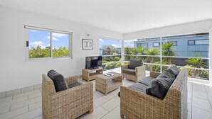 A modern living room with wicker furniture, a flat-screen TV, and large windows offering a view of greenery and a building. - Diamond Beach Beachfront Family Apt (Diamond beach)