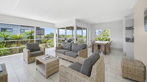 A spacious living area with wicker furniture, a dining table, and large windows offering a view of greenery. - Diamond Beach Beachfront Family Apt (Diamond beach)