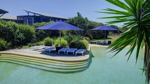 A pool area with lounge chairs and umbrellas, surrounded by greenery and a modern building in the background. - Diamond Beach Beachfront Family Apt (Diamond beach)