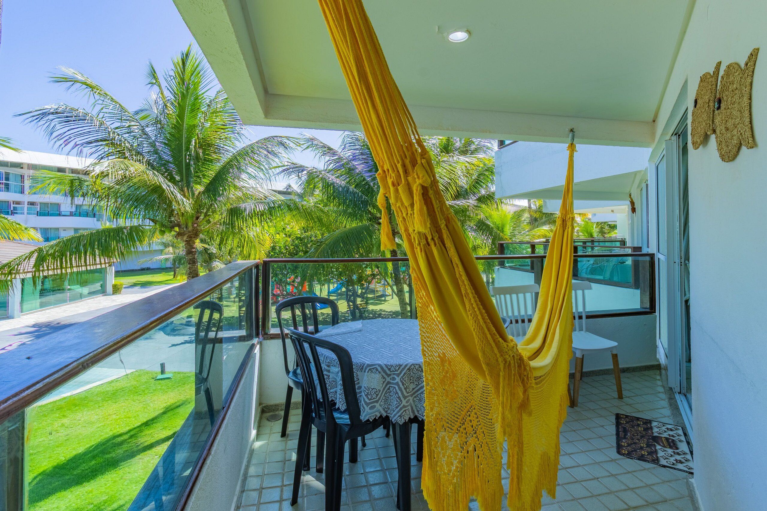 A balcony with a yellow hammock, a table with chairs, and a view of palm trees and buildings.
