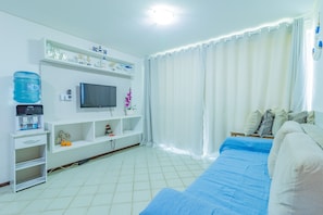 A bedroom with a bed, a TV on a stand, a water dispenser, and a shelf with decorative items. - Ancorar Resort #3103 - Beira Mar by Carpediem (Ipojuca)