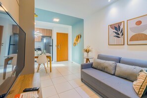 A modern living room with a grey sofa, a wooden coffee table, and a TV. There is a kitchen in the background with wooden cabinets and a fridge. - Mana Beach #d322 - Two Bedrooms by Carpediem (Ipojuca)