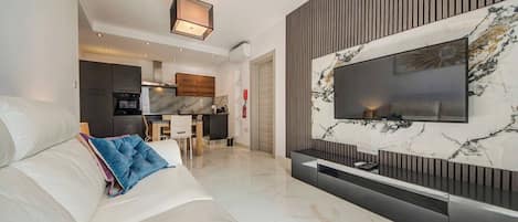 A modern living room with a large flat-screen TV mounted on a marble wall, a sofa with cushions, and a kitchen area in the background.