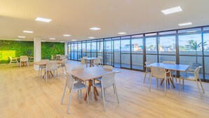 A spacious indoor area with multiple tables and chairs, large windows with a view of buildings and palm trees, and a green wall with plants. - Ora Beach #315 - Porto de Galinhas by Carpediem (Ipojuca)