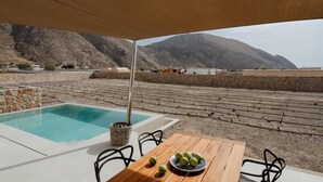 A poolside area with a wooden table and chairs, a stone wall, and a view of mountains and a body of water. - Rizes Villas Santorini with private pool (Santorini)