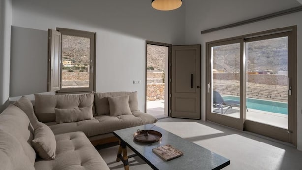 A modern living room with a beige sectional sofa, a coffee table, and a large sliding glass door leading to a pool area. - Rizes Villas Santorini with private pool (Santorini)